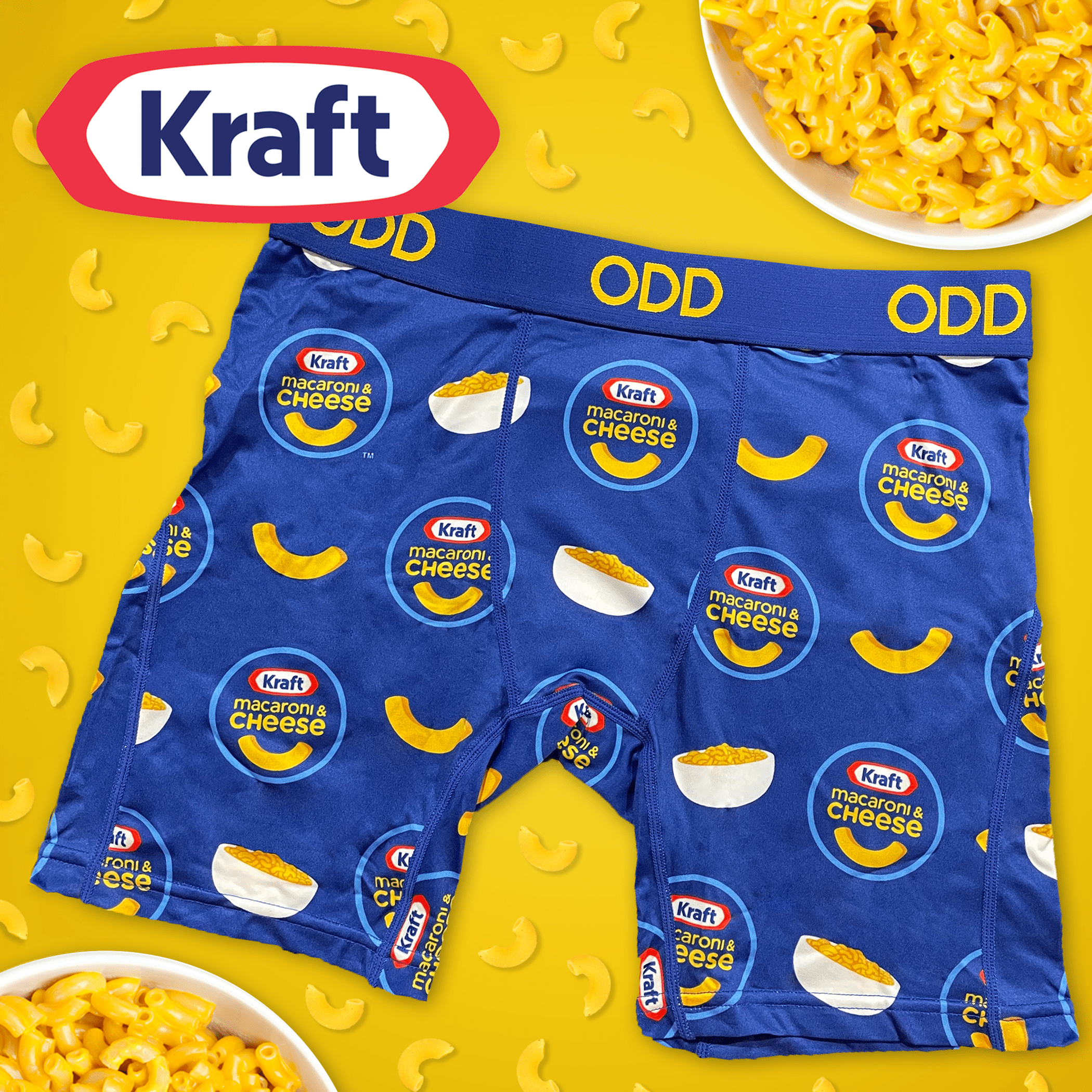 Blue boxer briefs featuring Kraft Macaroni & Cheese branding with logos, macaroni shapes, and bowls of mac and cheese. The waistband is blue with "ODD" branding in yellow, set against a yellow background scattered with macaroni.