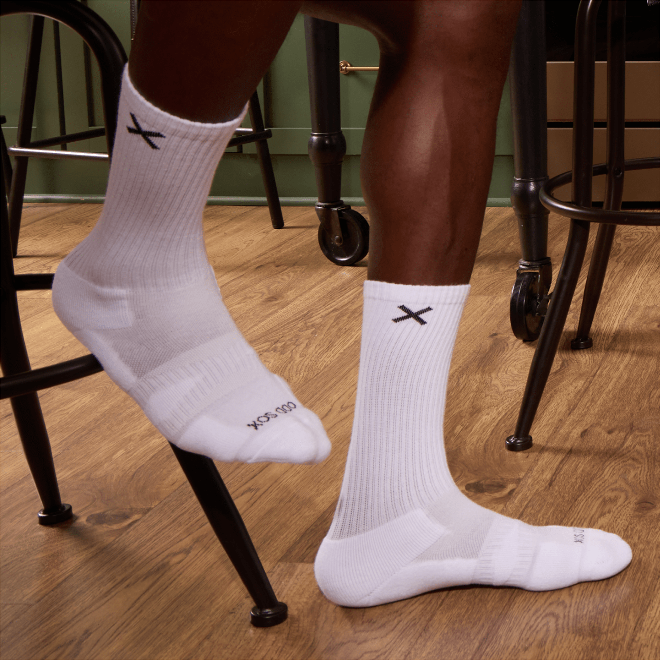 Man wearing white Basix crew socks with ribbed design and black X logo, seated at barstool.