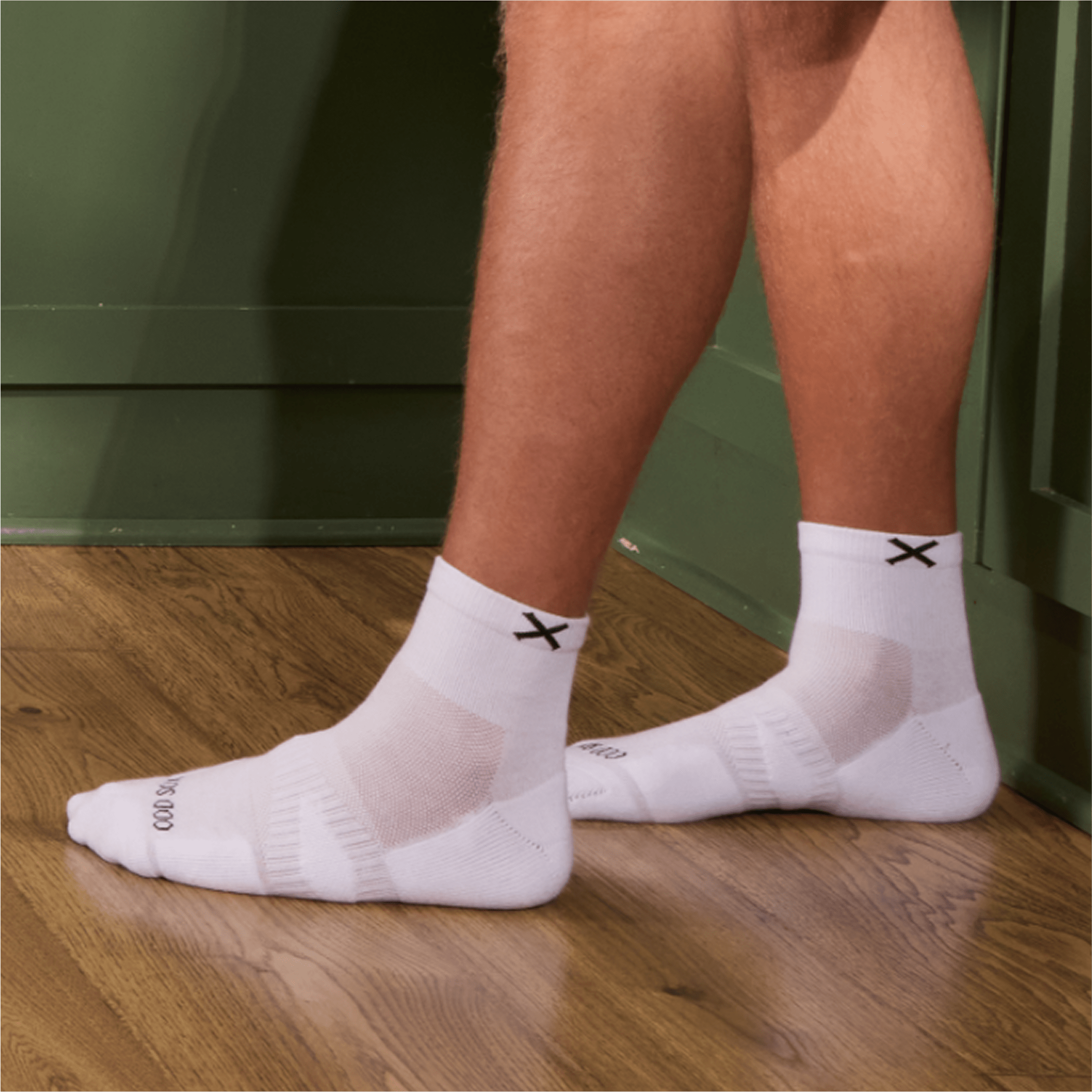 Man wearing white Basix quarter socks with black X logo while standing indoors.