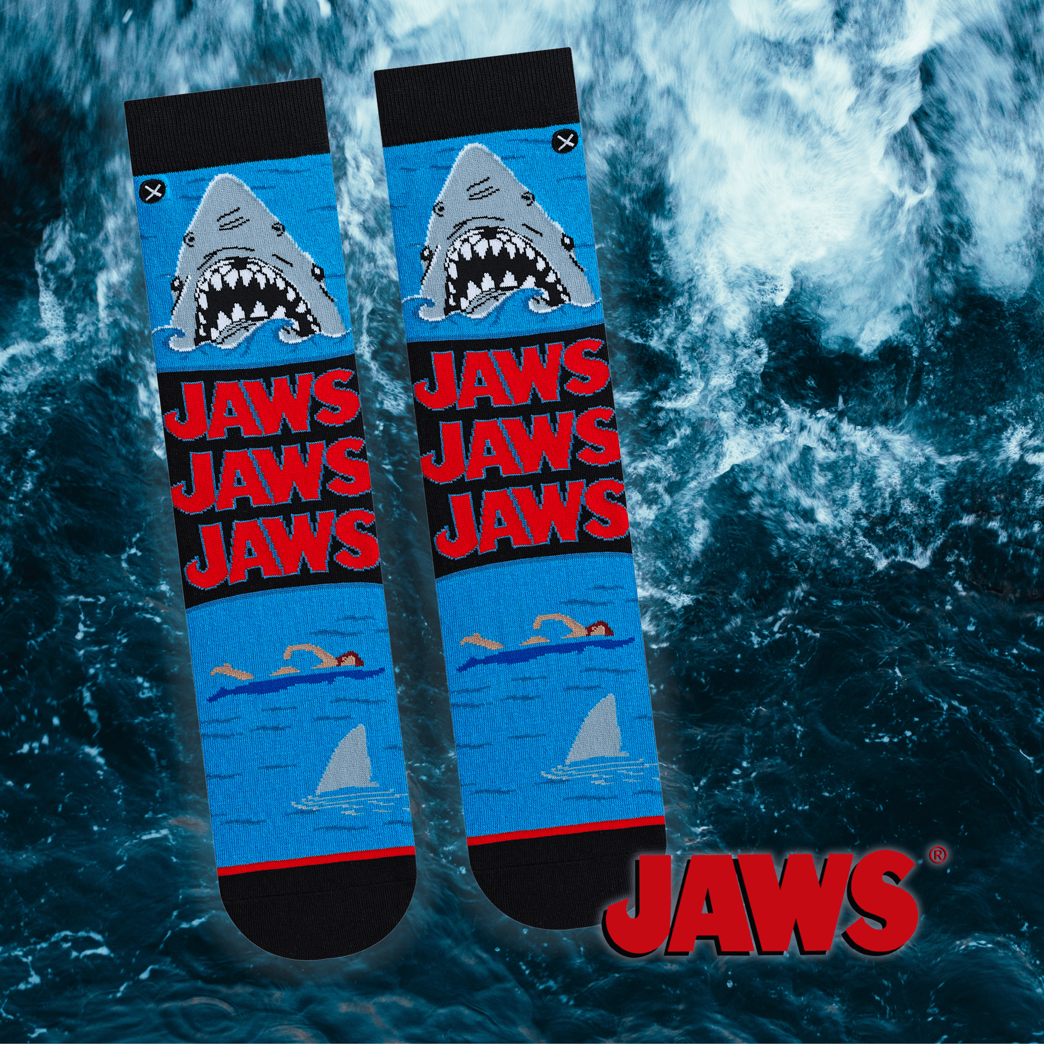 A pair of blue socks featuring the iconic shark from Jaws with the text "JAWS" repeated in bold red. Background shows ocean waves with the official Jaws logo.