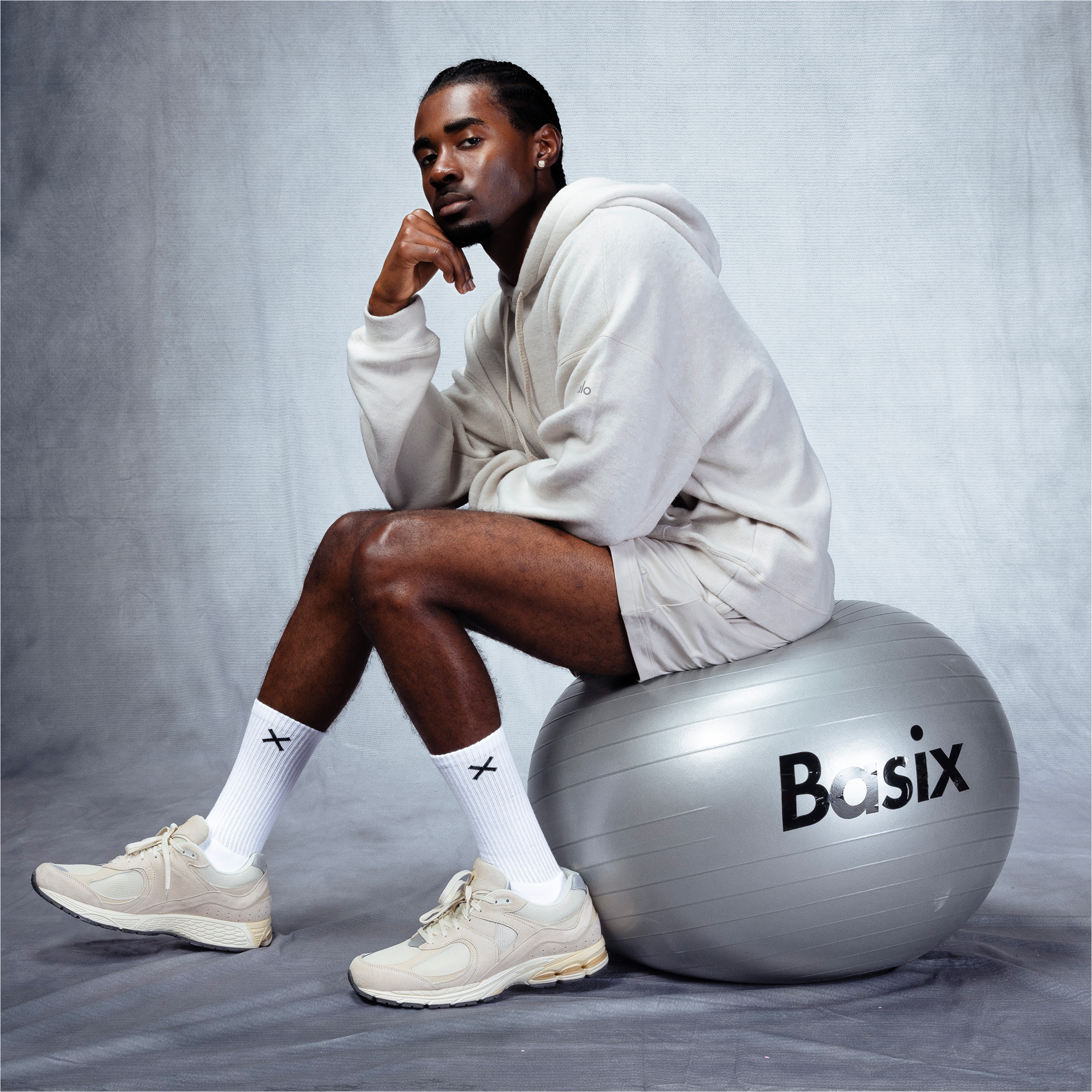 Male model in neutral hoodie and shorts sitting on a silver Basix exercise ball, wearing Basix white crew socks and sneakers for men’s collection.