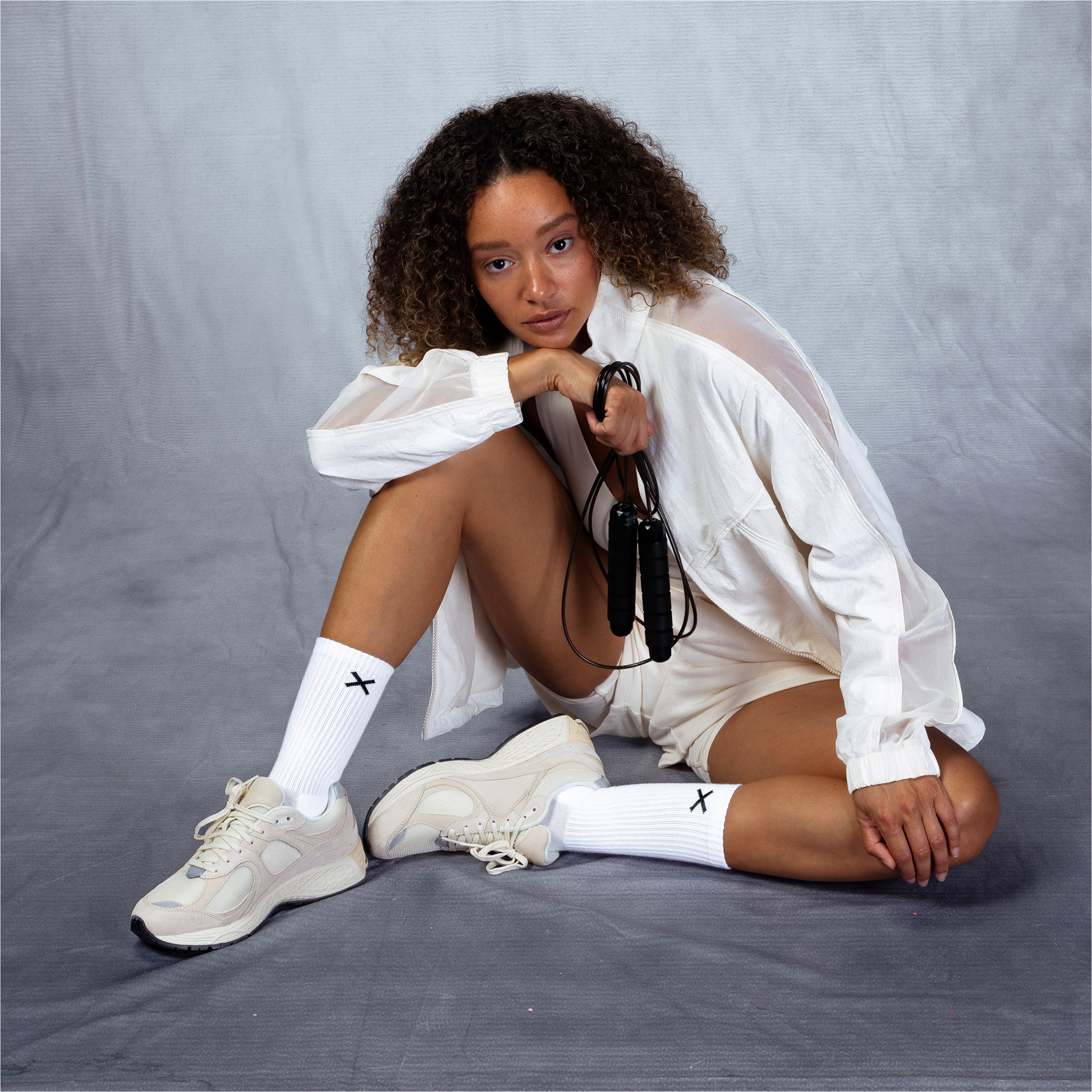 Female model in white activewear sitting on the floor holding a jump rope, styled with Basix white crew socks and sneakers for women’s collection.