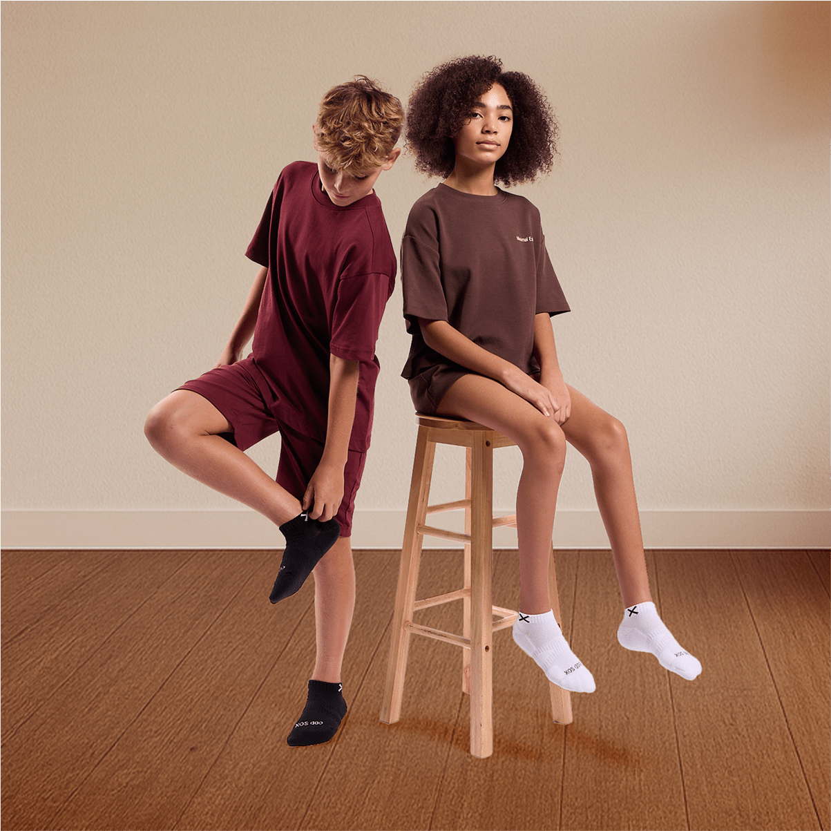 Two kids in matching burgundy loungewear wearing Odd Sox Basix ankle socks, one in black and one in white.