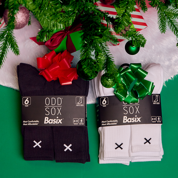 Holiday-themed image featuring two packs of Basix crew socks — one black and one white — placed under a Christmas tree with festive bows, ready for gifting.