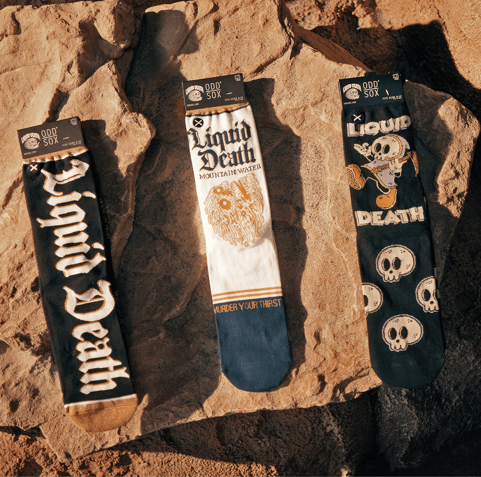 Three pairs of socks with 'Liquid Death' design on a rocky surface