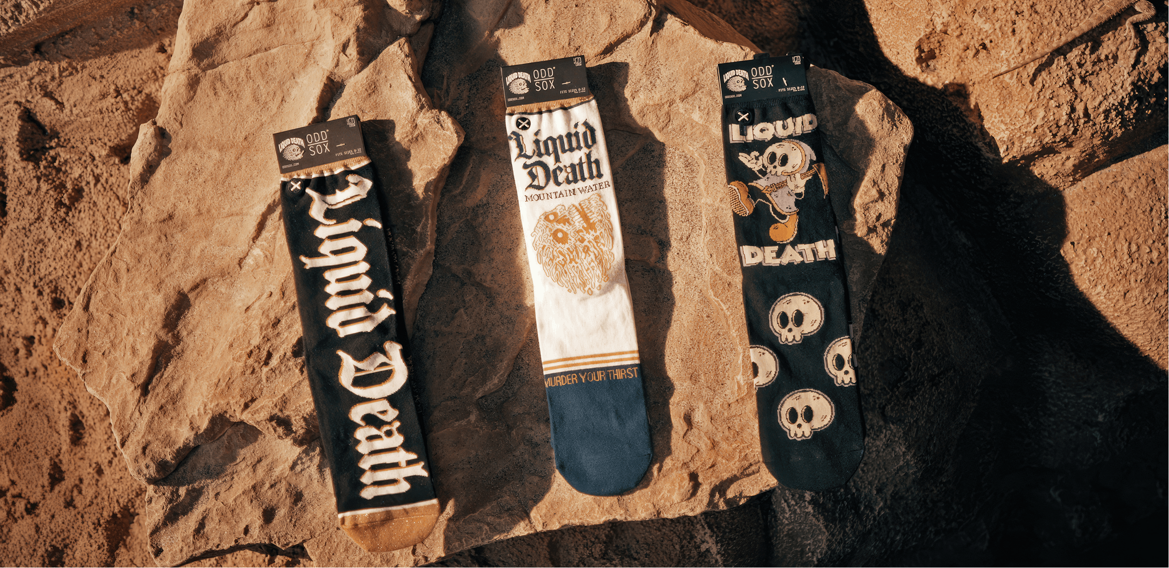 Three pairs of socks with 'Liquid Death' branding on a rocky surface