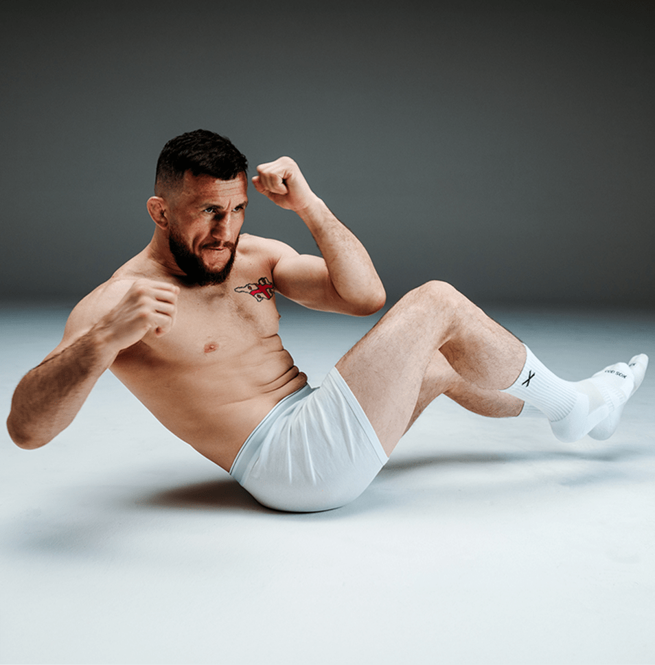 Man in white underwear sitting on a gray background, flexing his arm.