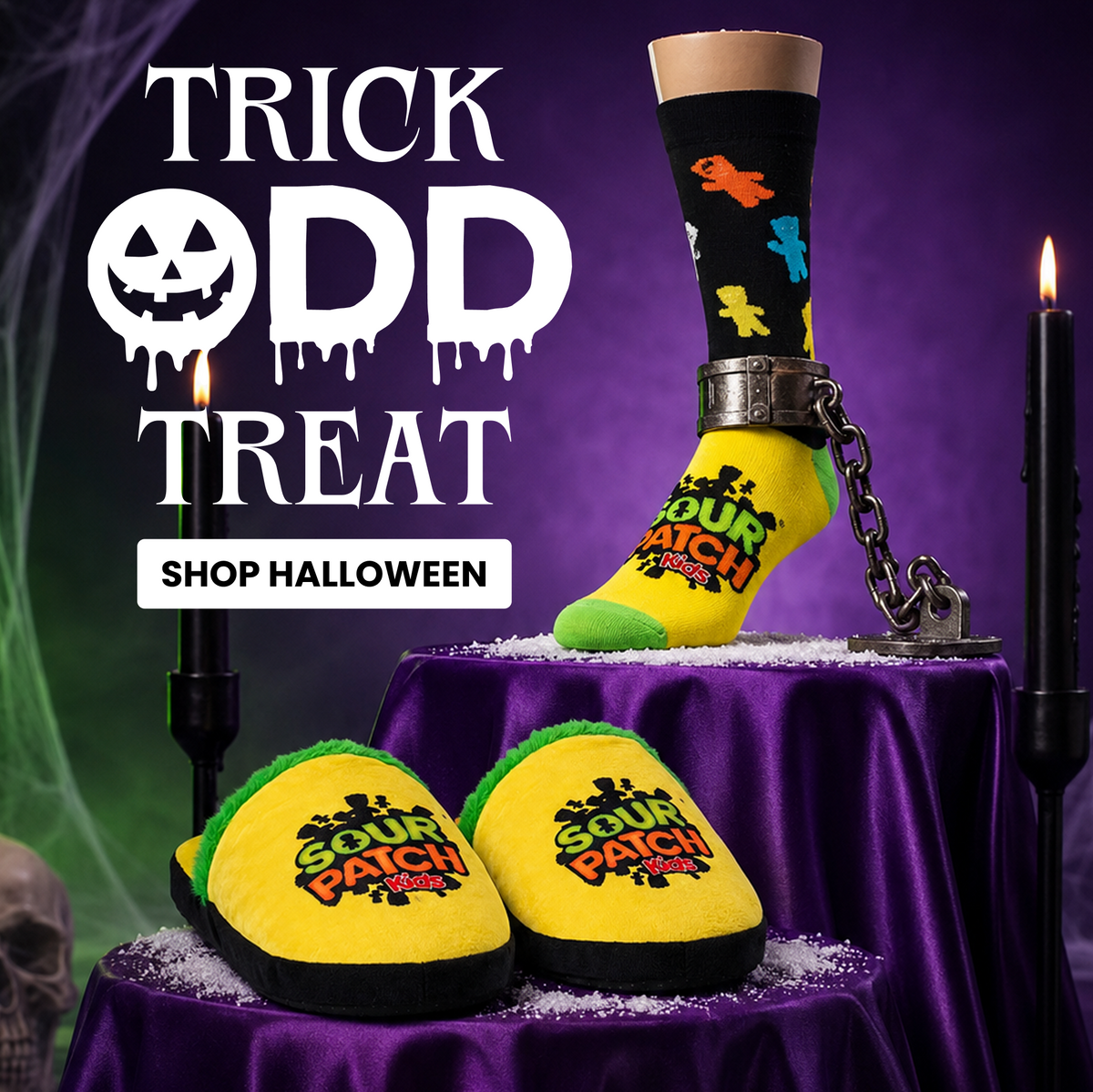 Halloween-themed product display featuring a pair of yellow and green 'Sour Patch' slippers on a purple draped pedestal, with a matching black sock on a leg prop. Large text reads 'TRICK OR TREAT' and 'SHOP HALLOWEEN' against a dark purple spooky background with candles and a skull.
