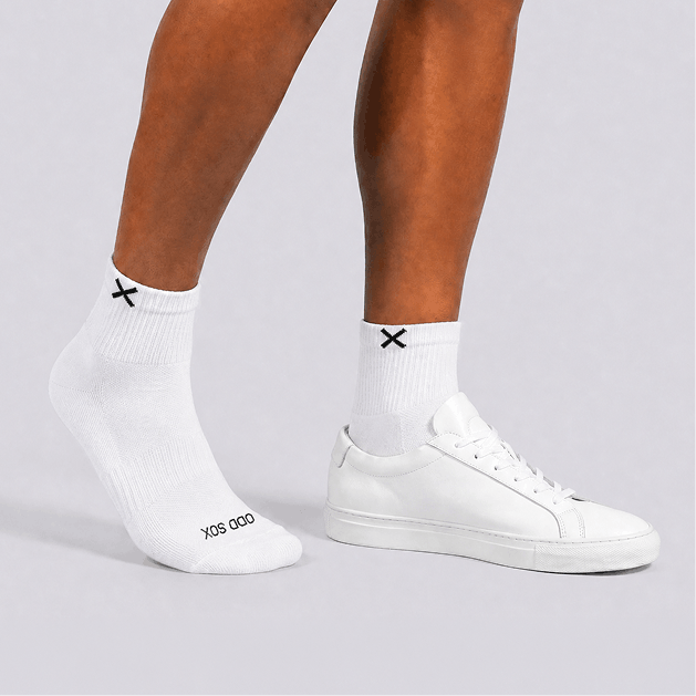 White socks with black 'ODD SOX' logo worn with white sneakers on a light gray background
