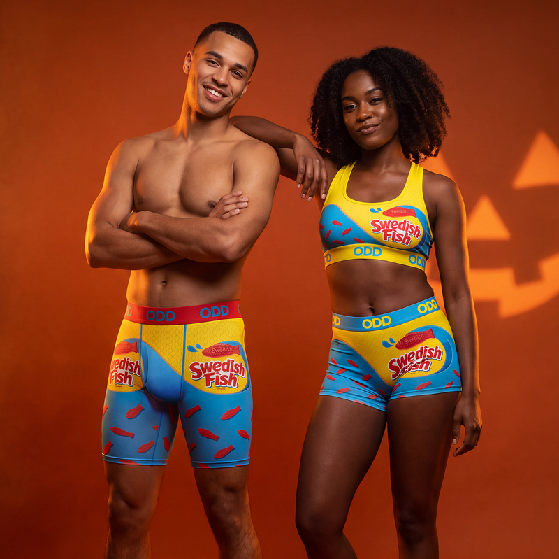 A shirtless man and a woman pose together against a warm orange studio background. The man wears colorful Swedish Fish branded boxer briefs, and the woman wears a matching yellow and blue sports bra with matching shorts.