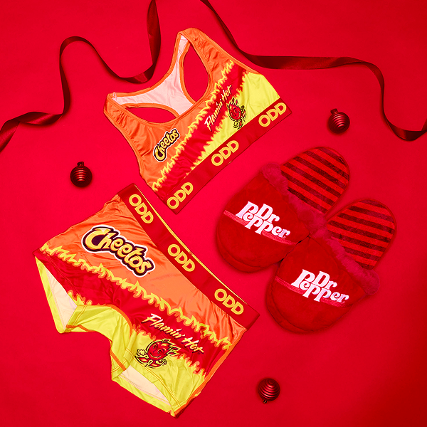 Festive holiday setup featuring women’s boy shorts, a Cheetos sports bra, and Dr Pepper slippers arranged together in a cozy, seasonal scene.